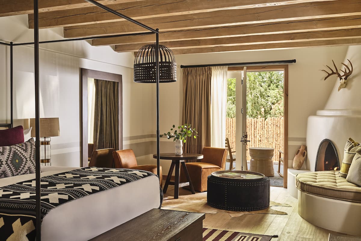 A well-lit room with a bed, a round ottoman, a chair, and a table.