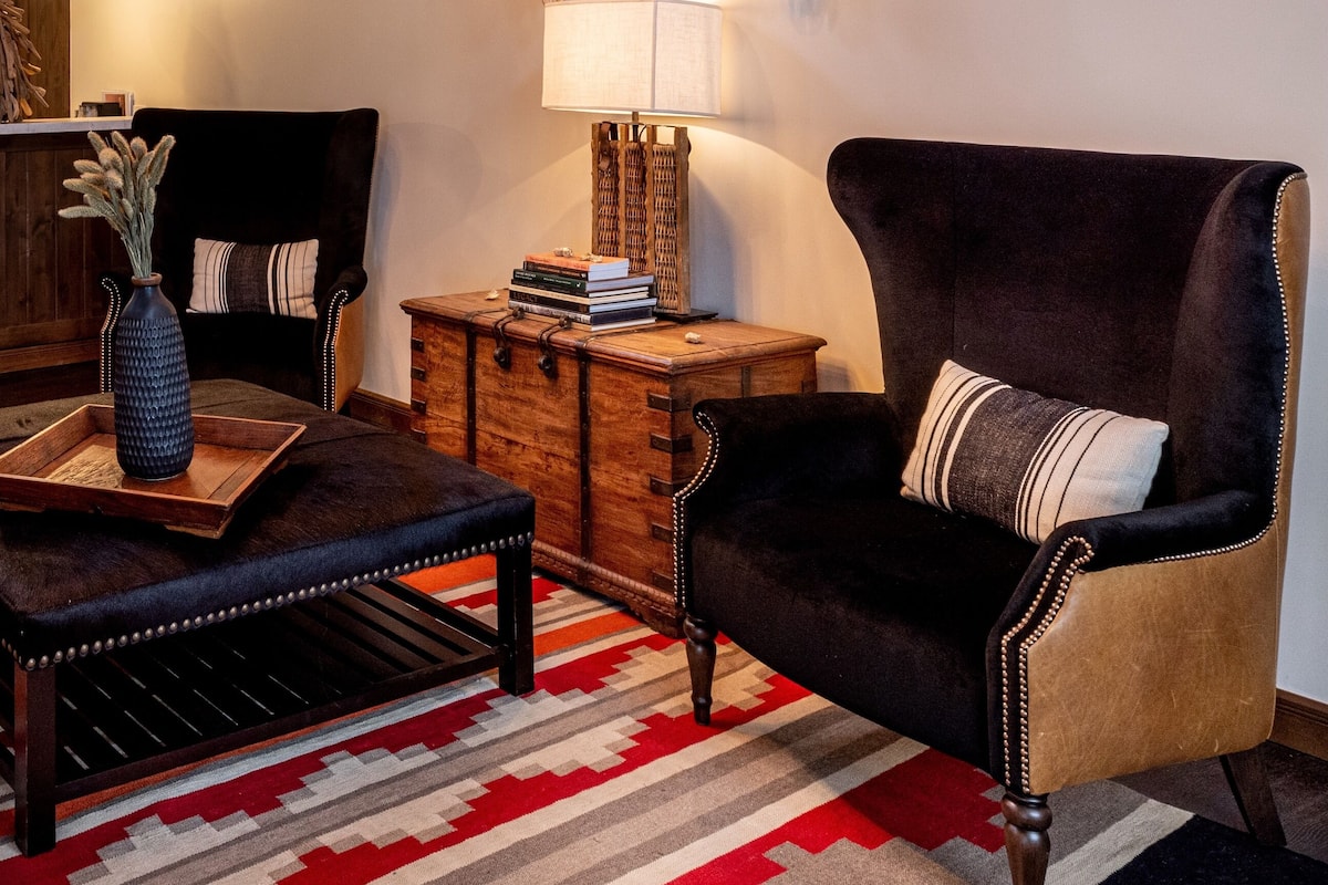 A room with a chandelier, a large wall-mounted skull display, a plush armchair, a wooden side table, and a patterned rug.
