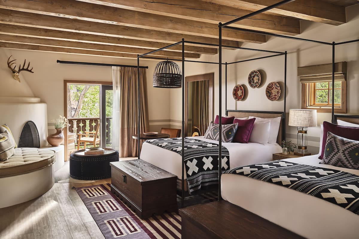 A four-poster bed with a patterned bedspread, a wooden bench, a small table, and a decorative wall piece.