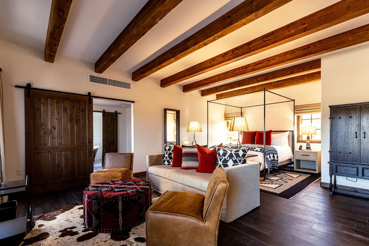 A spacious bedroom with a large bed, a sitting area with a sofa and chairs, wooden beams on the ceiling, and a wardrobe.