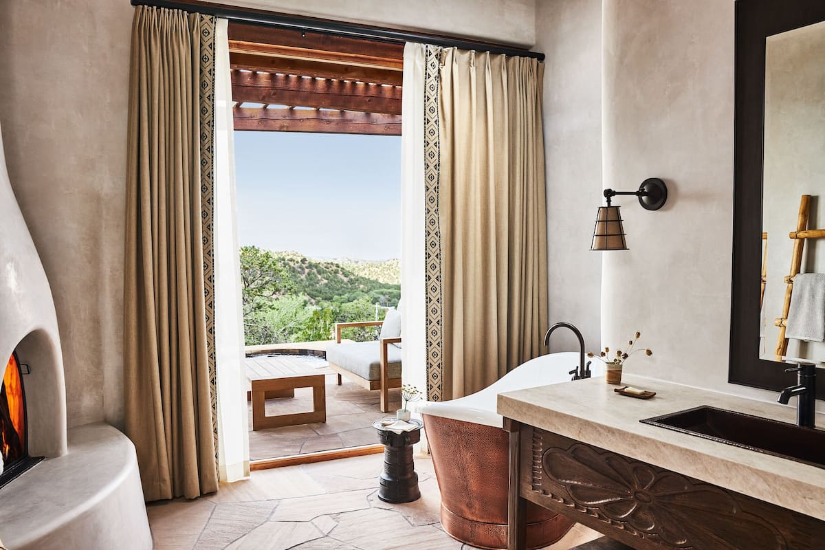 A bathroom with a bathtub, a sink, and a mirror. There is a window with curtains, a wooden bench, and a view of greenery outside.
