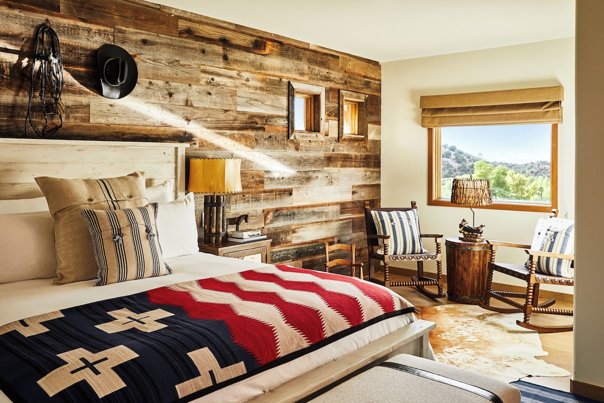 A bedroom with a wooden headboard, a large bed with a red and white striped blanket, a wooden rocking chair, and a view of the outdoors through a window.