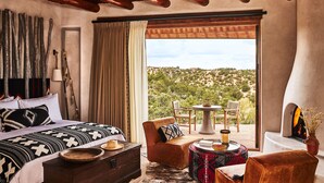 A bedroom with a bed, a sofa, a small table, and a fireplace. There is a view of a mountainous landscape through a large window. - Bishop's Lodge, Auberge Collection (Santa Fe)