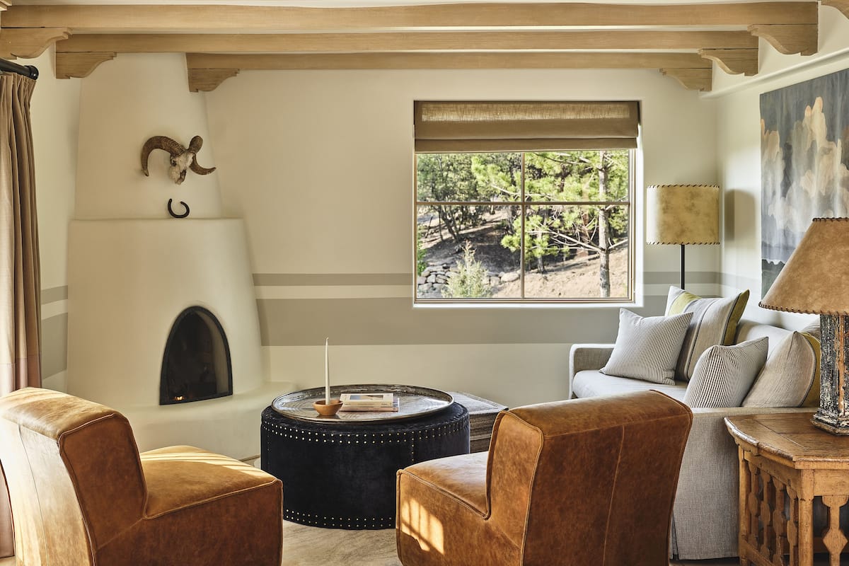 A cozy living room with a fireplace, leather armchairs, a round ottoman, and a large window with a view of trees.