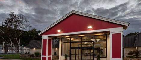 Un edificio con fachada roja y blanca, entrada de cristal y una zona de acceso marcado en el estacionamiento.