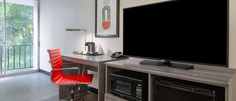 A modern office space with a large flat-screen TV, a red office chair, a desk with a lamp, a framed abstract artwork, and a built-in microwave.
