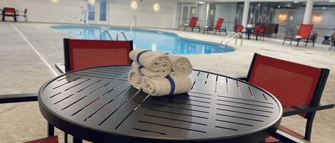Zona de piscina con una mesa redonda y sillas rojas, una pila de toallas y una piscina al fondo.