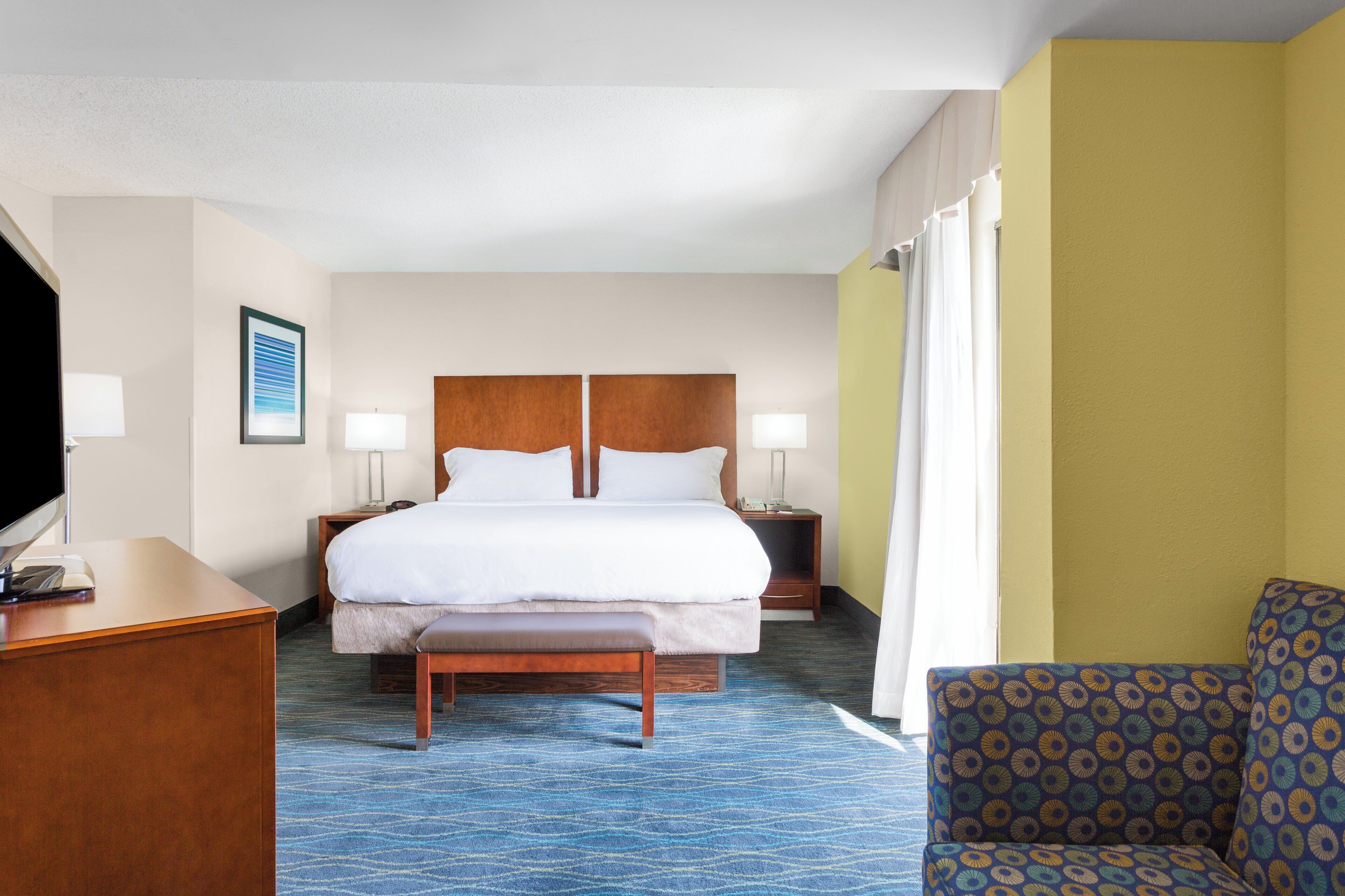 A hotel room with a bed, bedside tables, a desk, a chair, and a television.