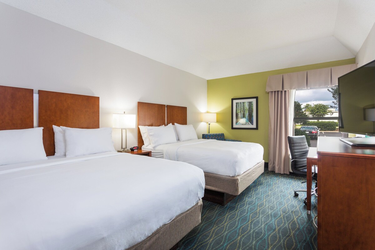 A hotel room with two beds, a desk, a chair, and a window with a view of the outdoors.