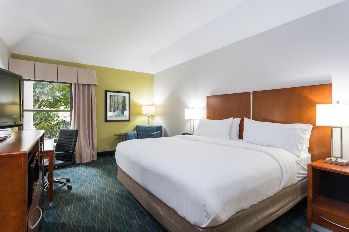 A hotel room with a large bed, a desk with a computer, a chair, a TV, and a window with curtains.