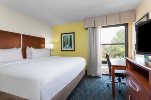 A hotel room with a large bed, a desk with a chair, a television, and a window with a view of trees.