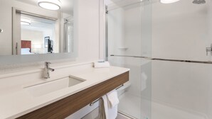 Un baño moderno con cabina de ducha de vidrio, un lavamanos blanco con tocador de madera y un espejo grande.