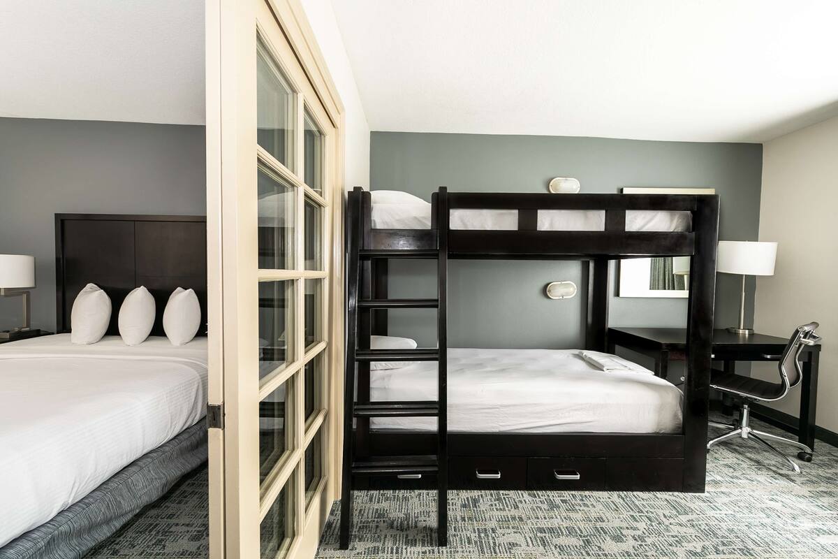 Habitació d'hotel amb un llit superposat, un escriptori amb una cadira, una làmpada i una finestra gran.