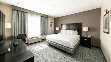 Quarto de hotel com uma cama grande, uma escrivaninha e uma cadeira. Há uma janela com cortinas, uma TV na parede e um quadro emoldurado.