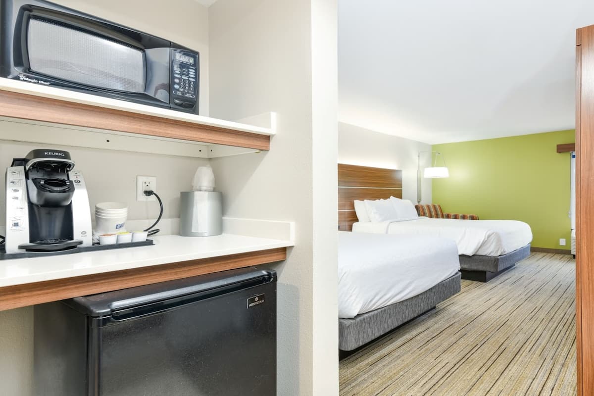 A kitchenette with a microwave, coffee maker, and refrigerator. It also features a bed with two pillows and a bedside table.