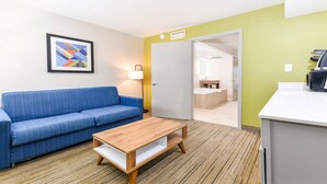Une chambre d’hôtel avec un canapé bleu, une table basse en bois, un tableau abstrait encadré, une lampe et une kitchenette équipée d’un évier et d’un plan de travail.