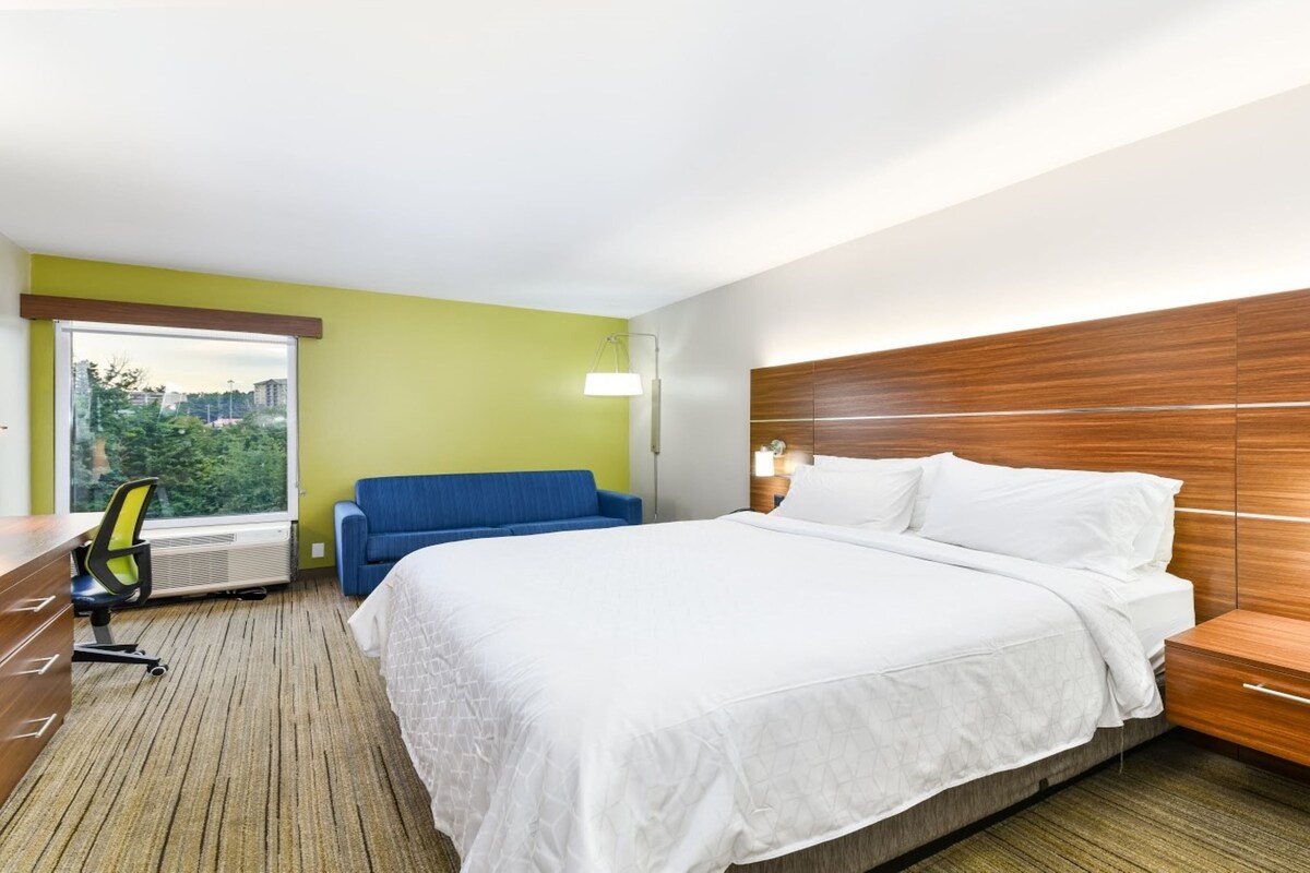 A hotel room with a large bed, a blue sofa, a desk with a chair, and a window with a view of trees and buildings.