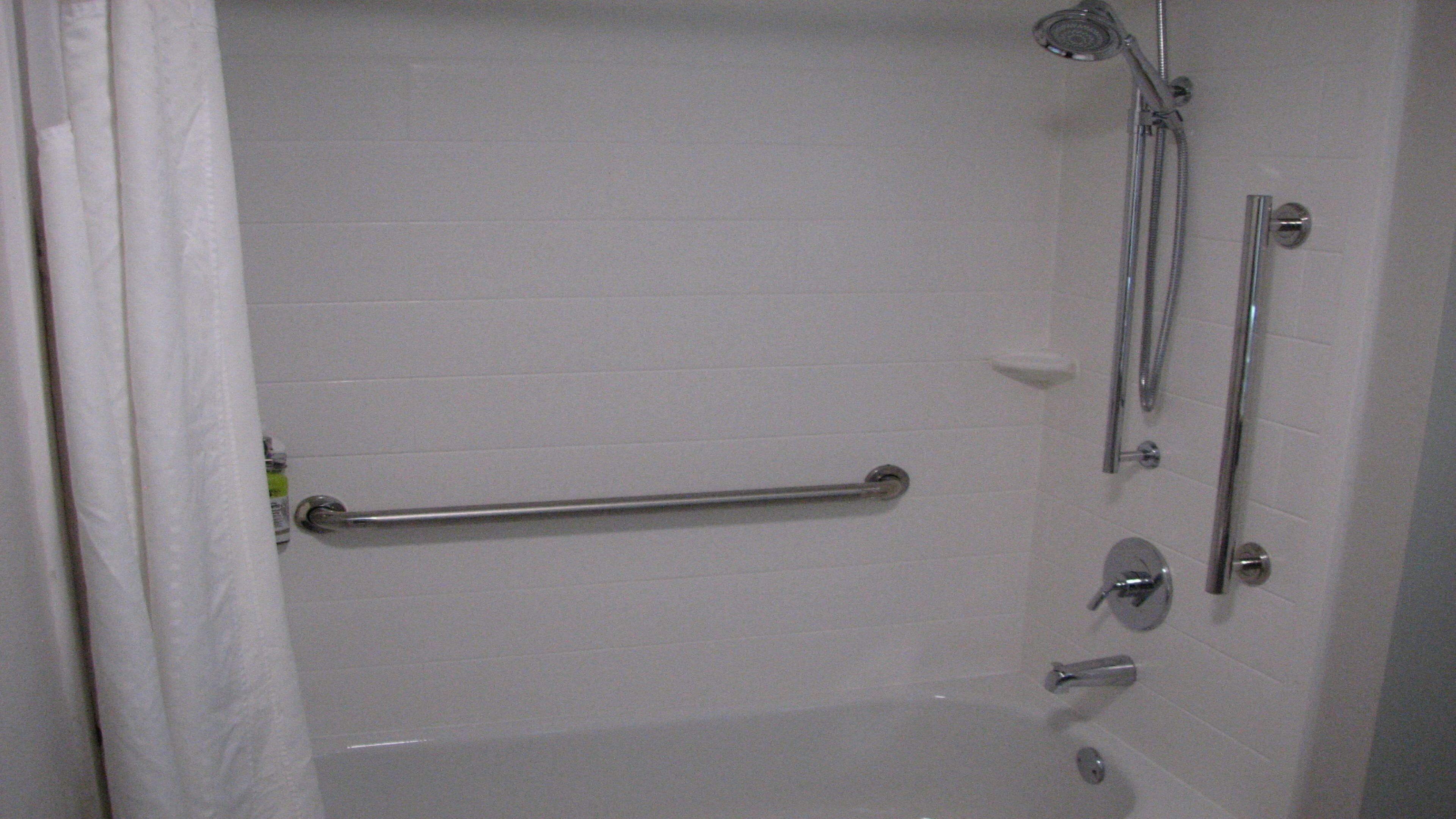 A bathroom with a white tiled wall, a shower with a handheld showerhead, a grab bar, and a toilet with a privacy partition.