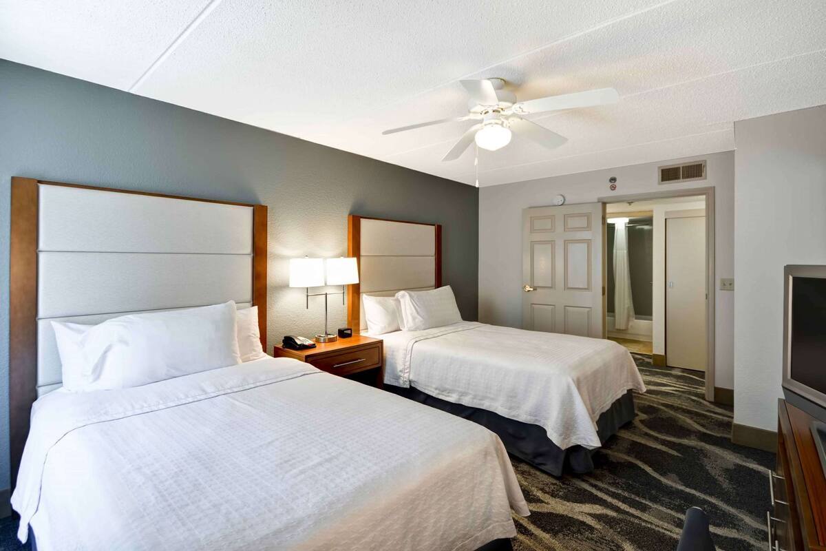 A hotel room with two beds, a ceiling fan, a TV, and a door leading to a bathroom.