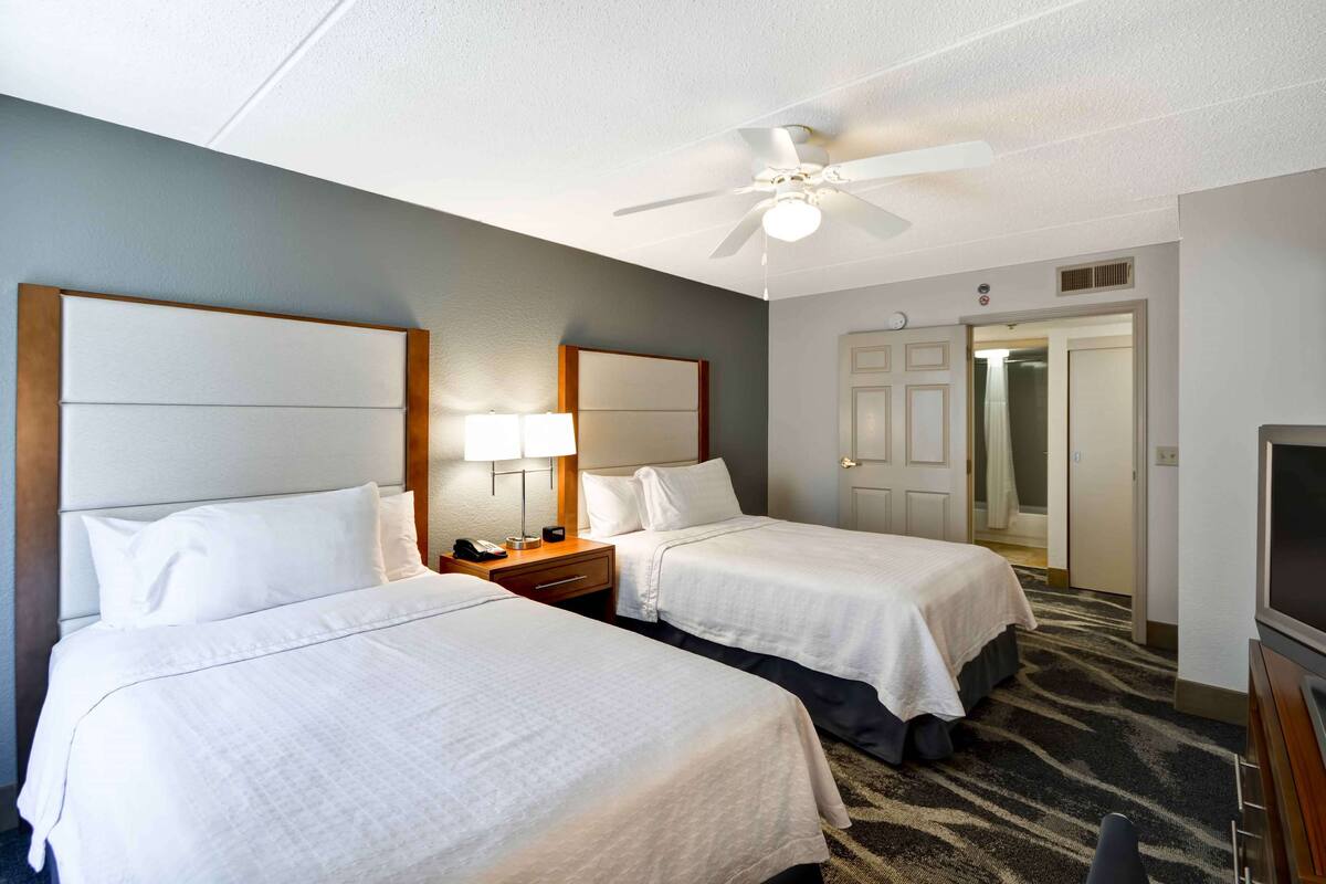 A hotel room with two beds, a ceiling fan, a TV, and a door leading to a bathroom.