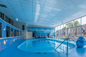 Una piscina interior con un mural de vida marina en las paredes, una zona de estar junto a la piscina con sillas y ventanas amplias que dejan entrar luz natural.