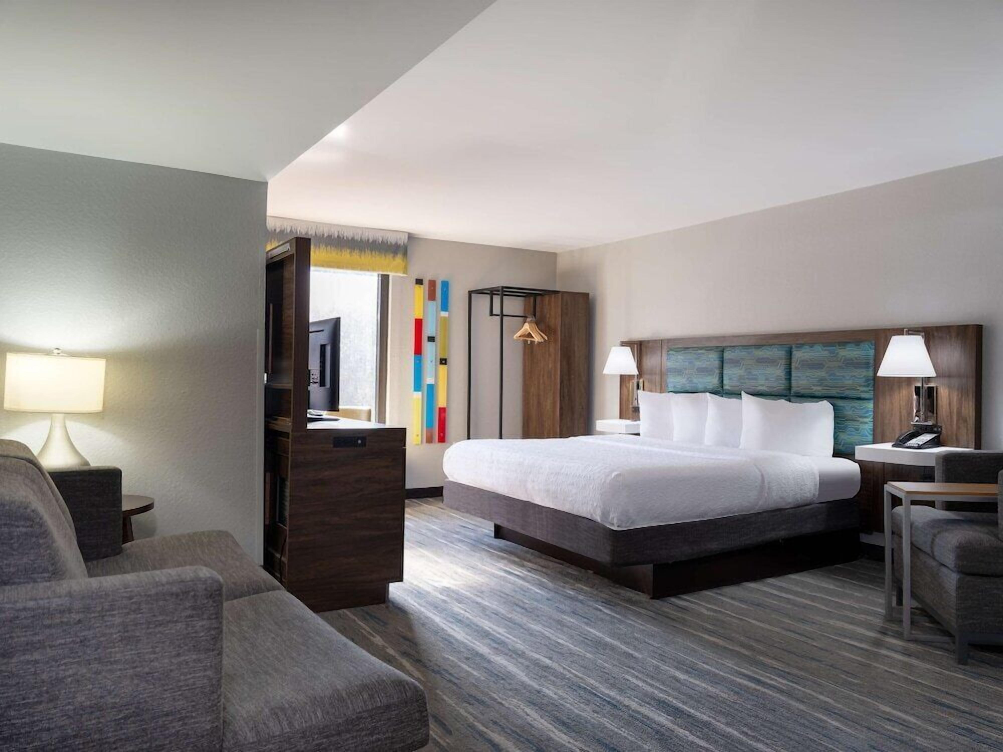 Quarto de hotel com cama, escrivaninha, cadeira, televisão e guarda-roupa.