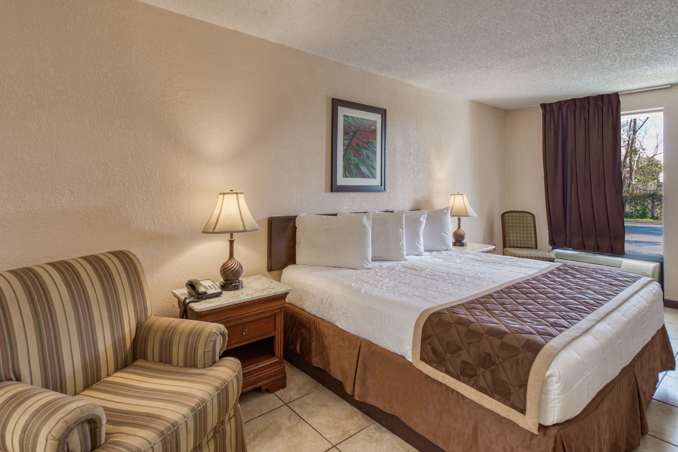 A hotel room with a bed, a striped armchair, a nightstand with a lamp, and a window with curtains.