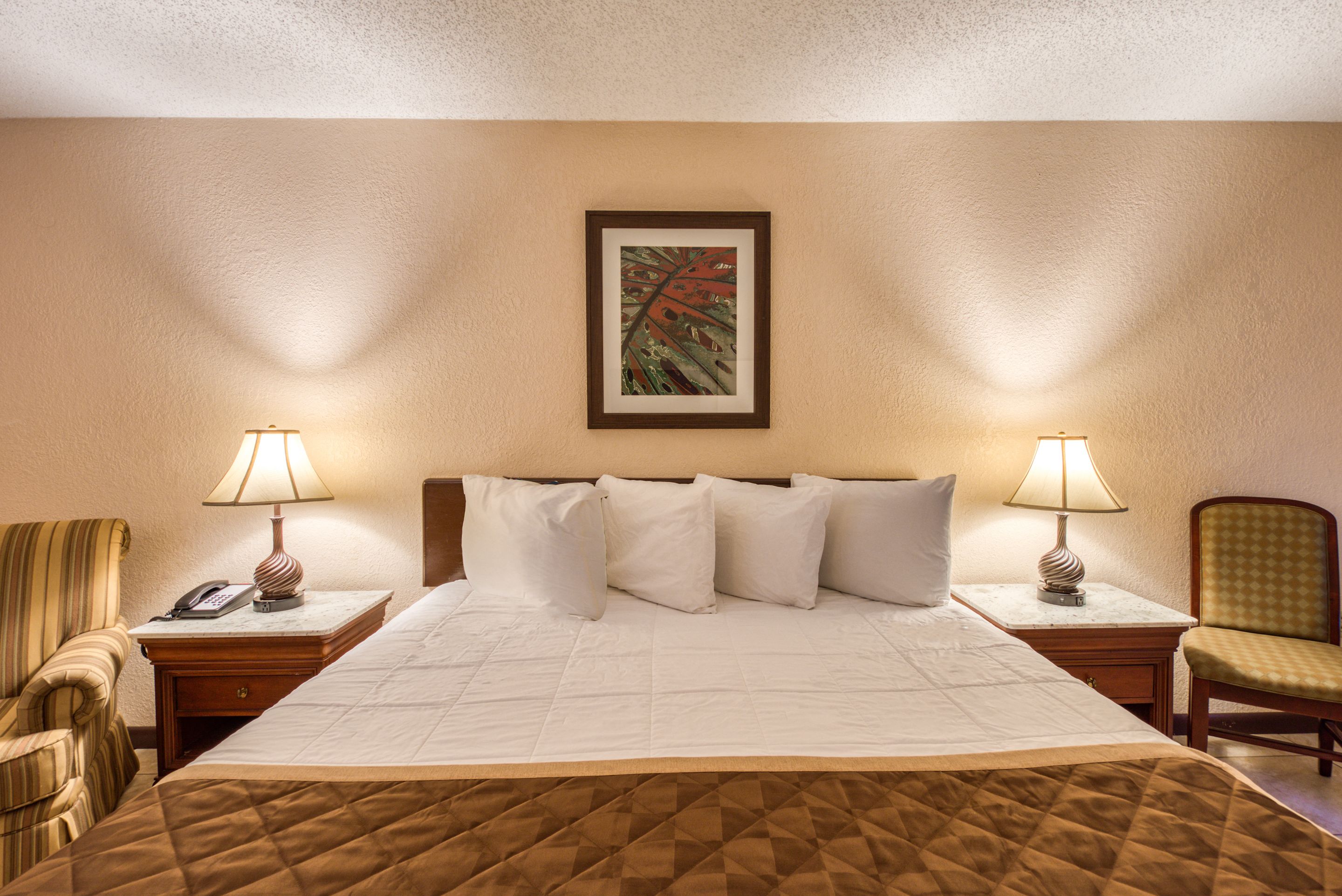 A hotel room with a large bed, two bedside tables with lamps, a chair, and a framed artwork on the wall.