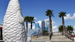 Una gran escultura de pez blanco junto a una piscina, con palmeras y edificios modernos al fondo.