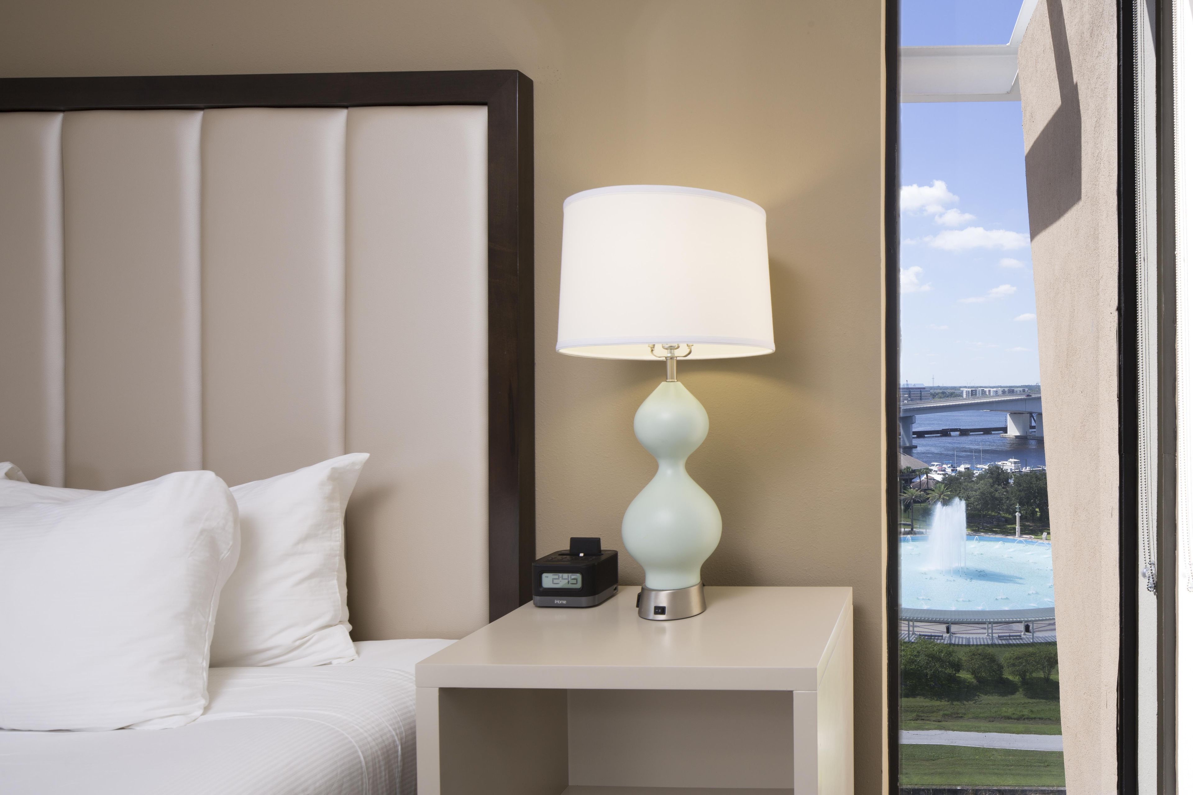 A hotel room with a bed, a nightstand with a lamp, and a view of a fountain and bridge.