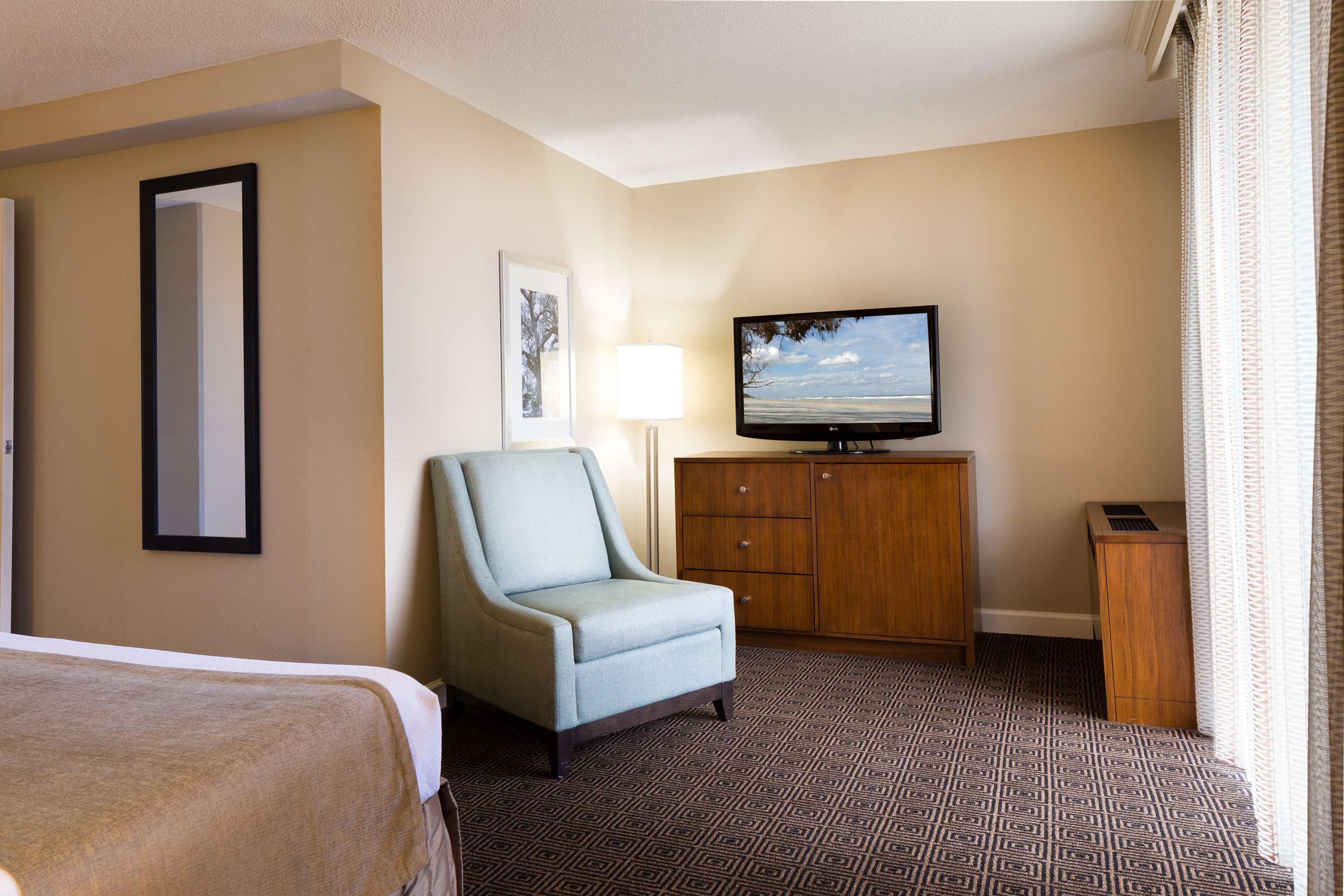 A hotel room with a bed, a light blue armchair, a wooden cabinet, a television, and a patterned carpet.