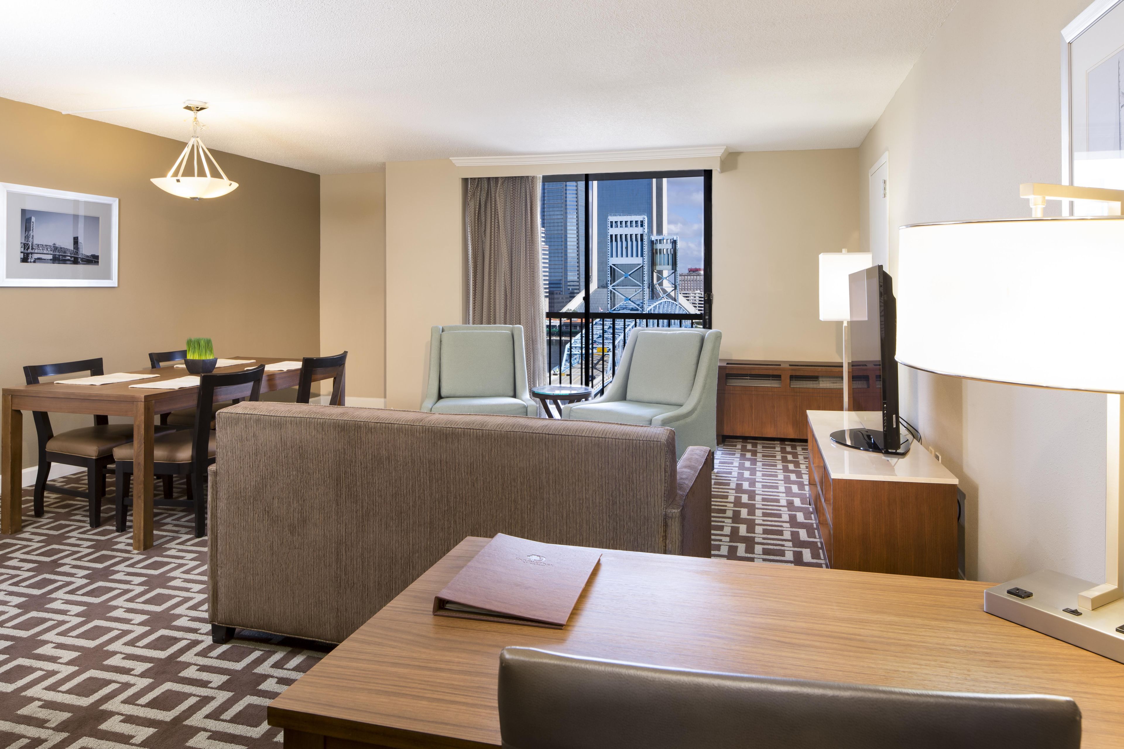 A hotel room with a dining area, two armchairs, a sofa, a table with a folder, a lamp, and a view of a bridge and buildings.