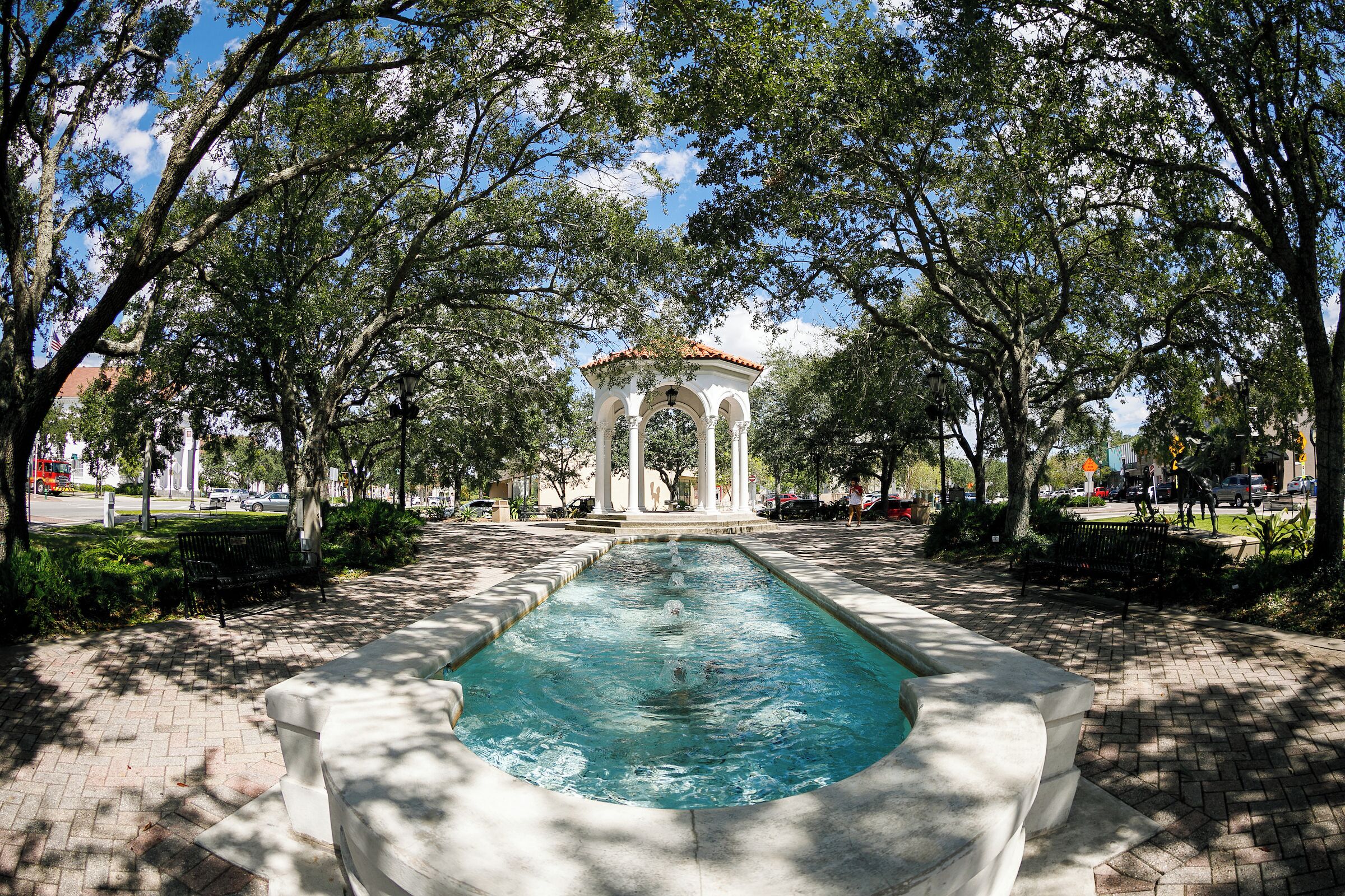 A rectangular pool with a white gazebo in the center, surrounded by trees and a paved walkway.