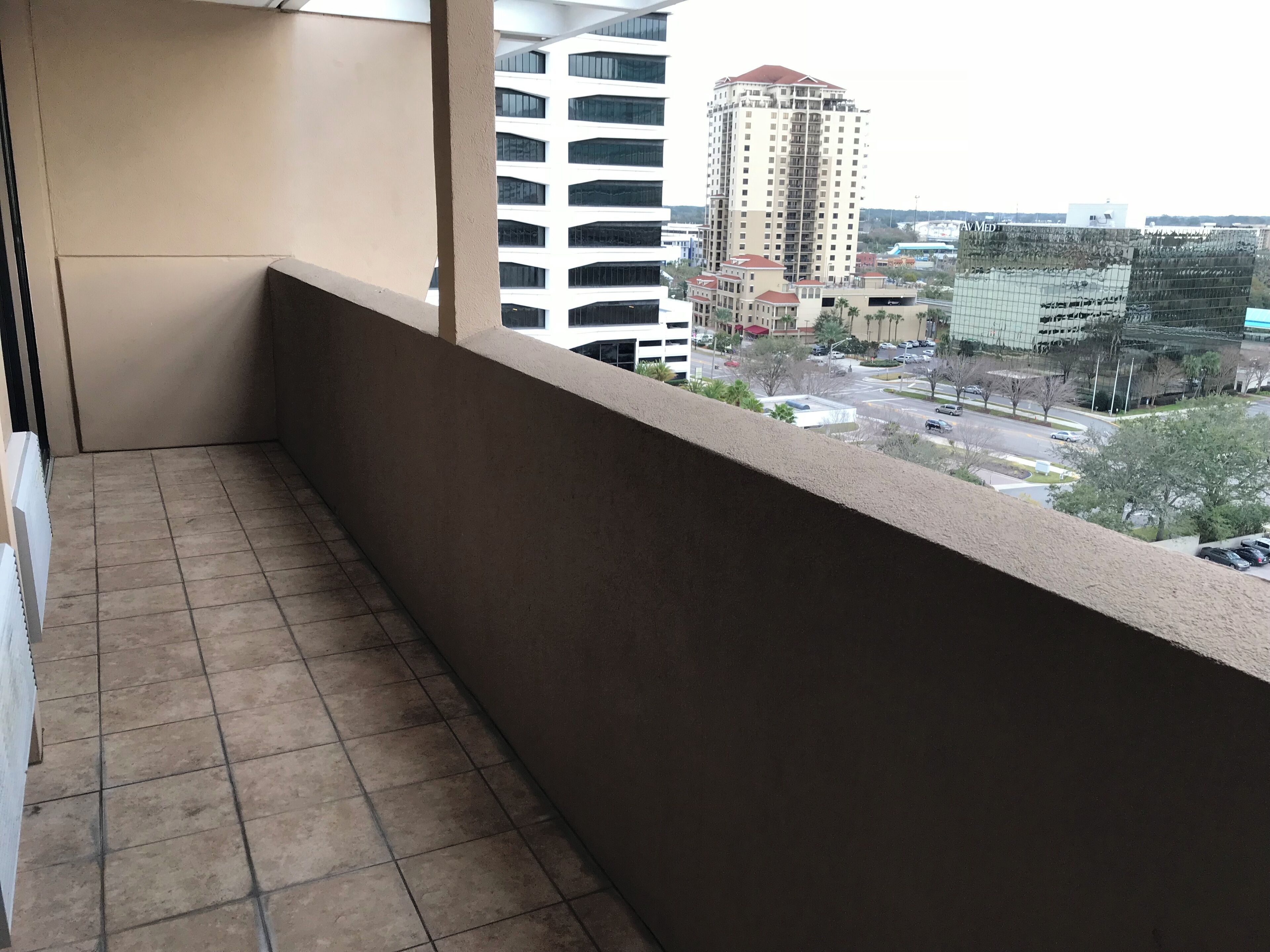 Un balcó amb sòl de rajola i vista a un paisatge urbà amb edificis alts i una zona d'aparcament.