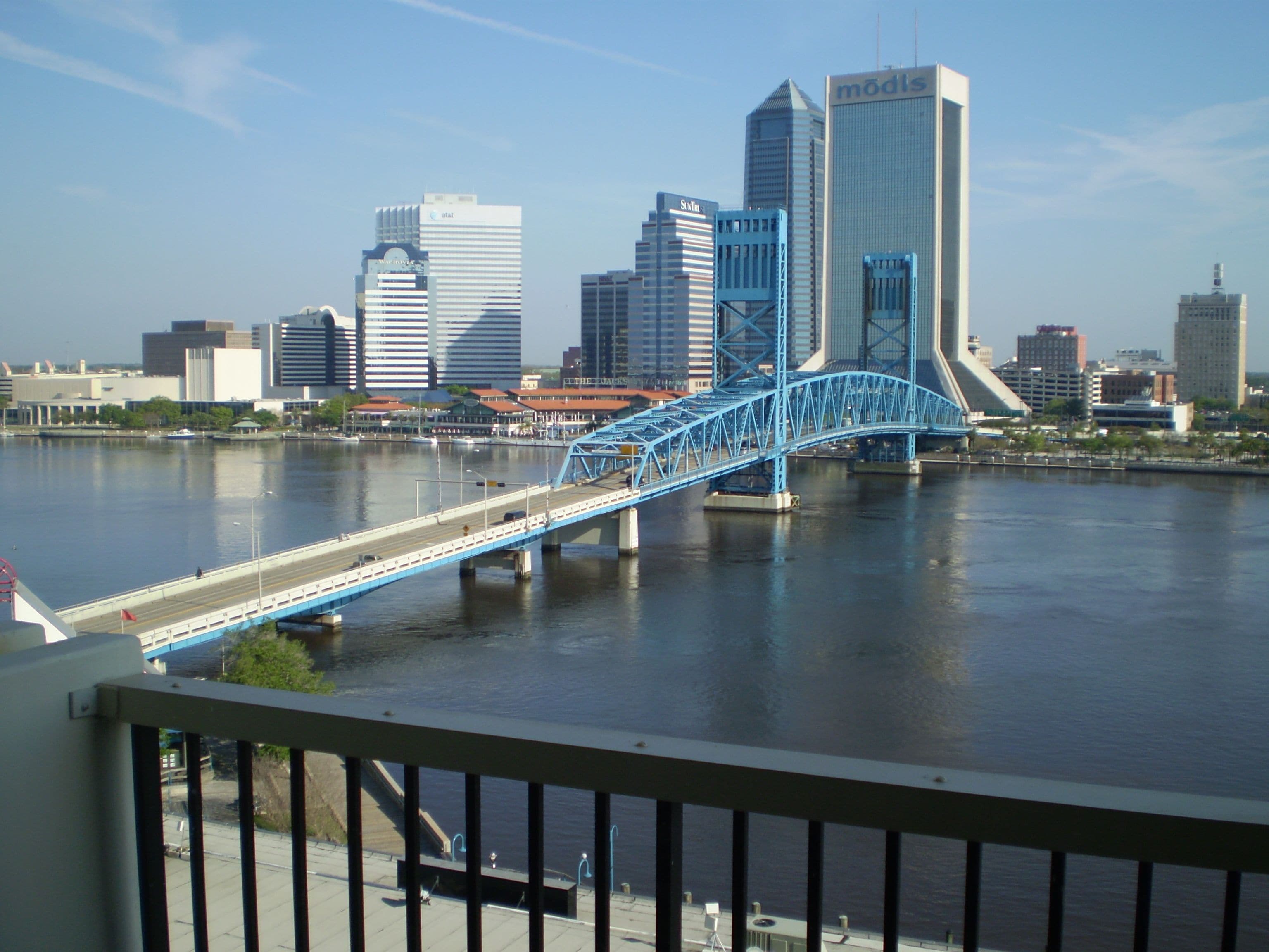 Un paisaje urbano con un puente azul destacado sobre un río, edificios modernos y un cielo despejado.
