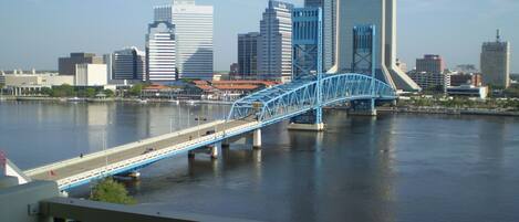 Un paisaje urbano con un puente azul destacado sobre un río, edificios modernos y un cielo despejado.
