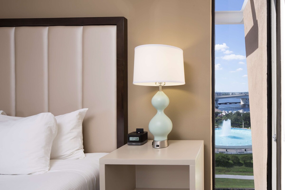 Een hotelkamer met een bed, een nachtkastje met een lamp, een digitale klok en uitzicht op een fontein door een raam.