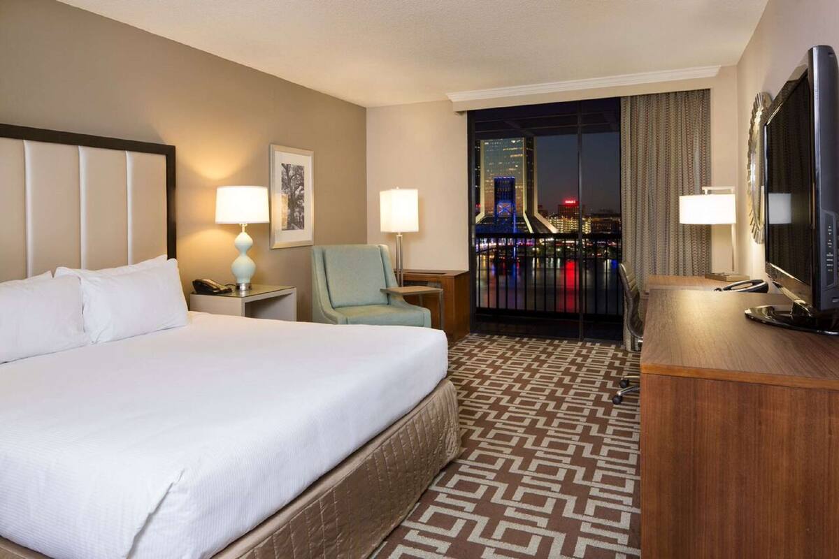 Une chambre d’hôtel avec un grand lit, deux lampes de chevet, une chaise, un bureau avec une télévision et une vue sur le paysage urbain la nuit.