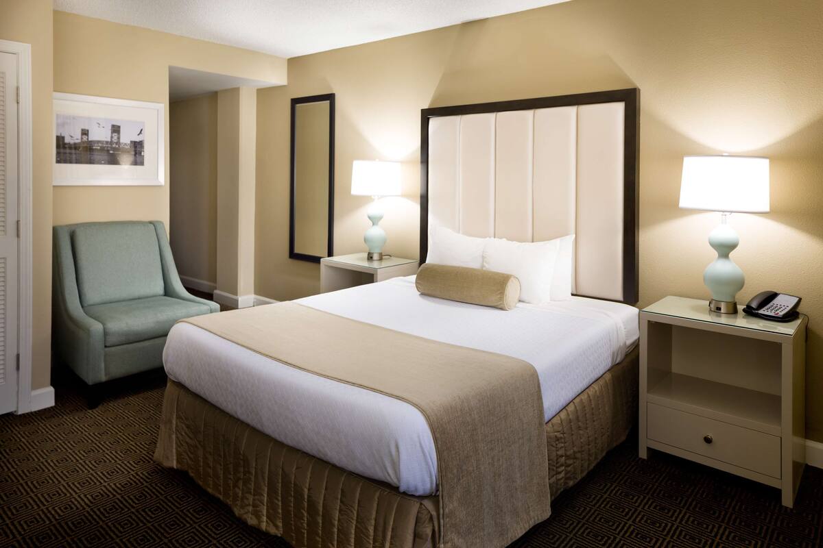 Une chambre d’hôtel avec un grand lit, deux tables de chevet avec des lampes, une chaise, un miroir et un tableau encadré au mur.