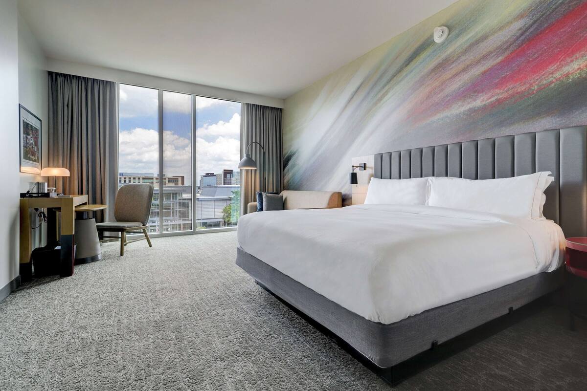 A hotel room with a large bed, a desk, a chair, and a view of the city through large windows.
