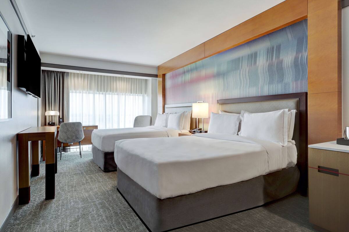 A hotel room with two beds, a desk, a chair, a TV, and a large abstract painting on the wall.