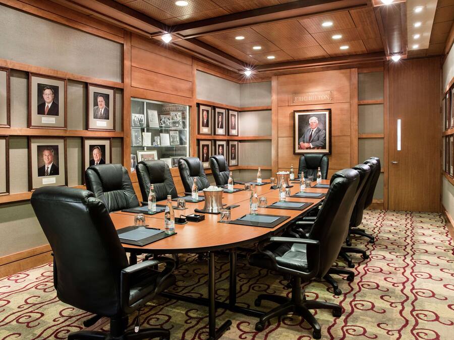 A conference room with a long table, black leather chairs, framed pictures on the walls, and a patterned carpet.