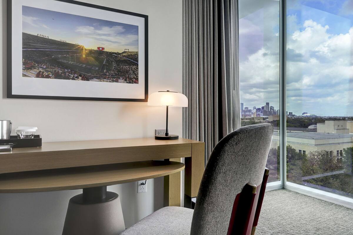 A hotel room with a large window, a desk, a chair, a framed picture, and a lamp.