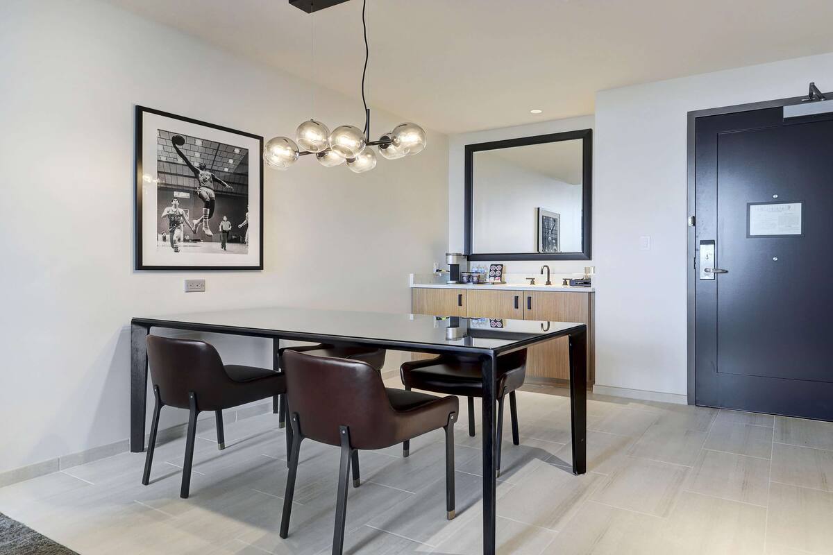 A modern dining area with a black dining table, four chairs, a pendant light, a framed picture on the wall, a kitchen area with a sink and cabinets, and a dark door with a sign.