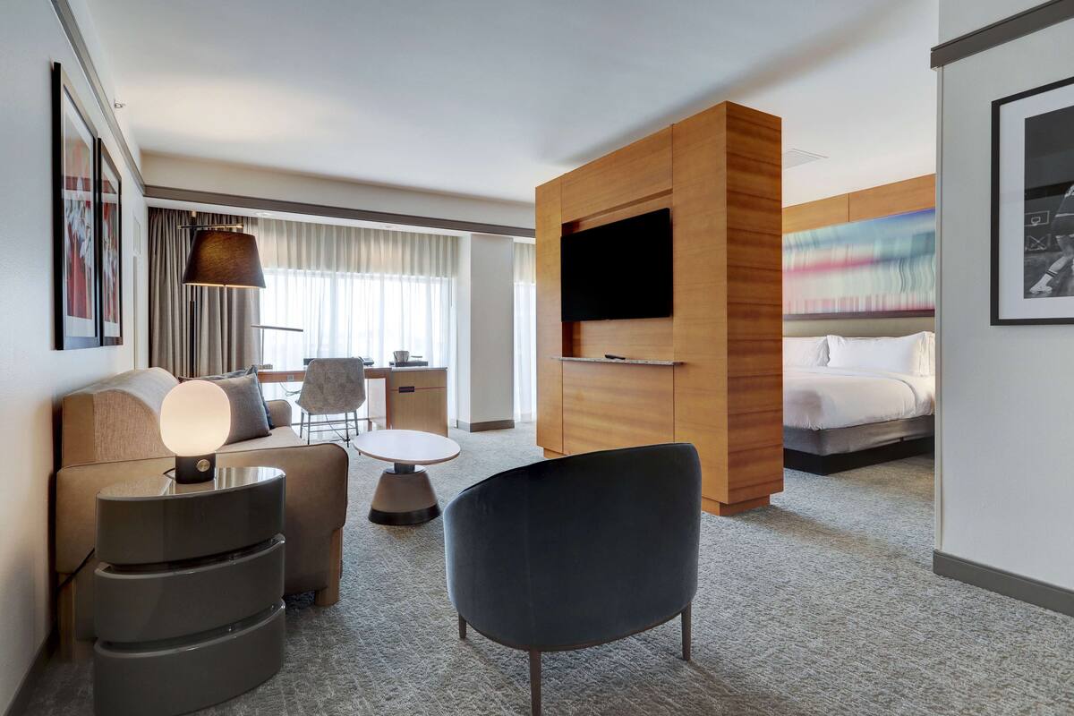 A modern hotel room with a sofa, a chair, a small table, a TV mounted on a wooden panel, and a bed with a white comforter.