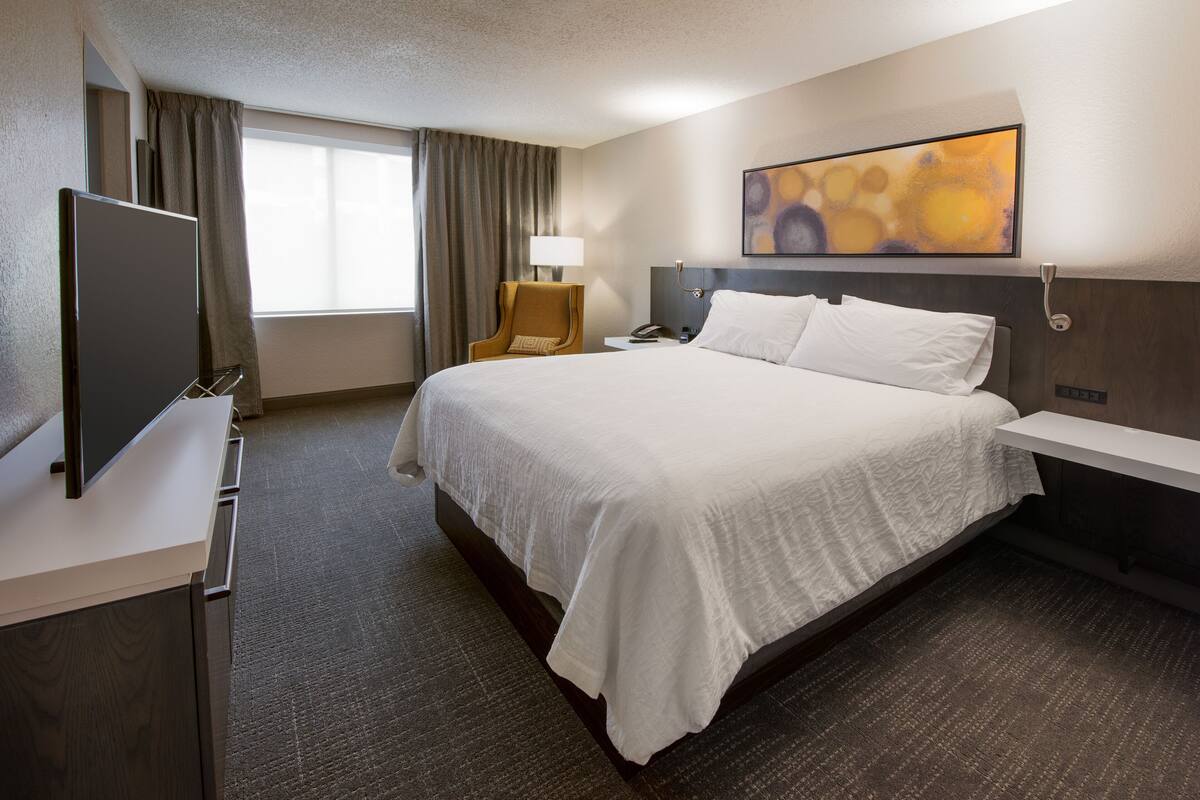 Una habitación de hotel con una cama grande, un escritorio con televisión de pantalla plana, una silla, una mesita de noche con lámpara y un cuadro en la pared.