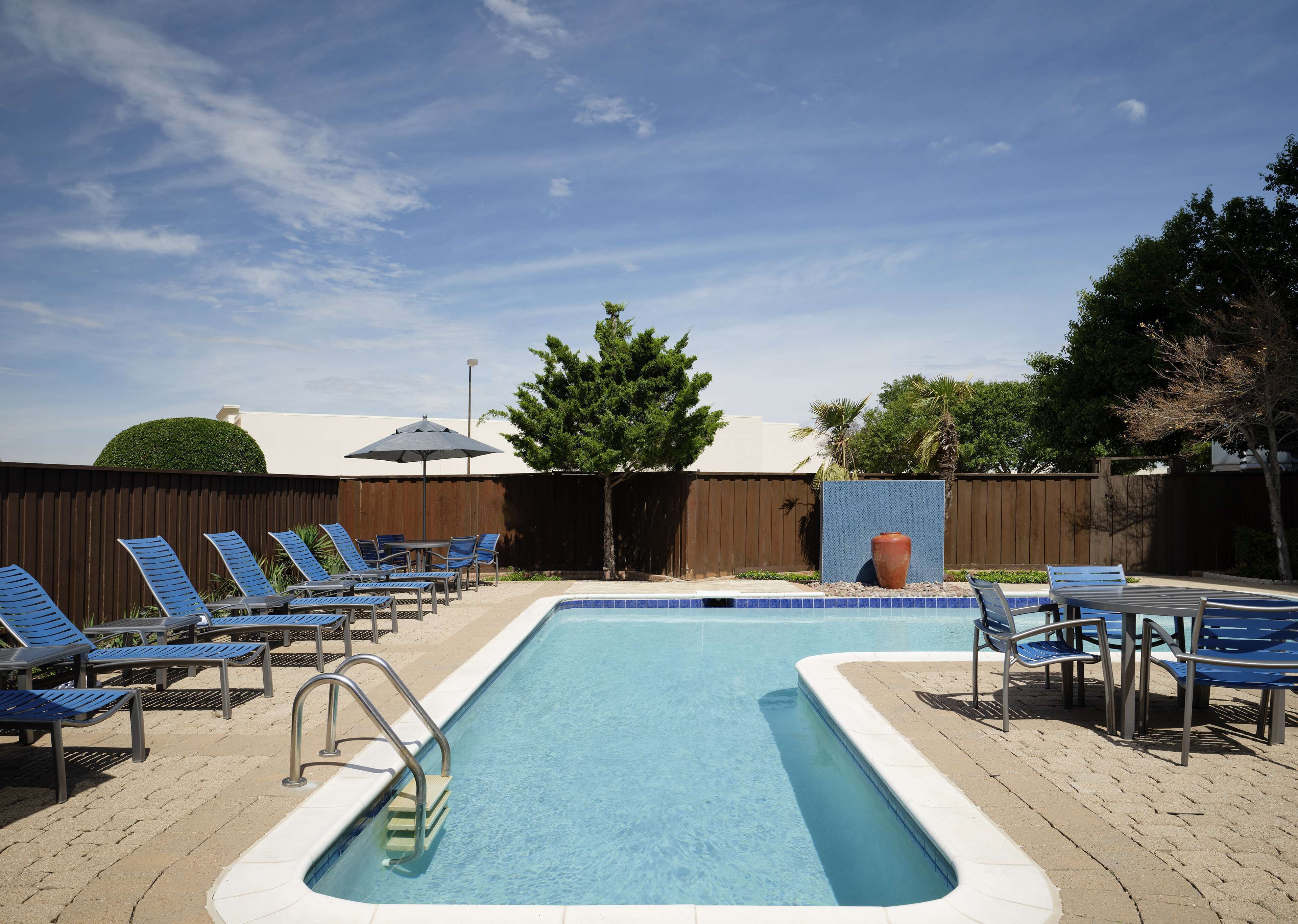 A rectangular swimming pool with a tiled edge, surrounded by lounge chairs and a table with an umbrella.