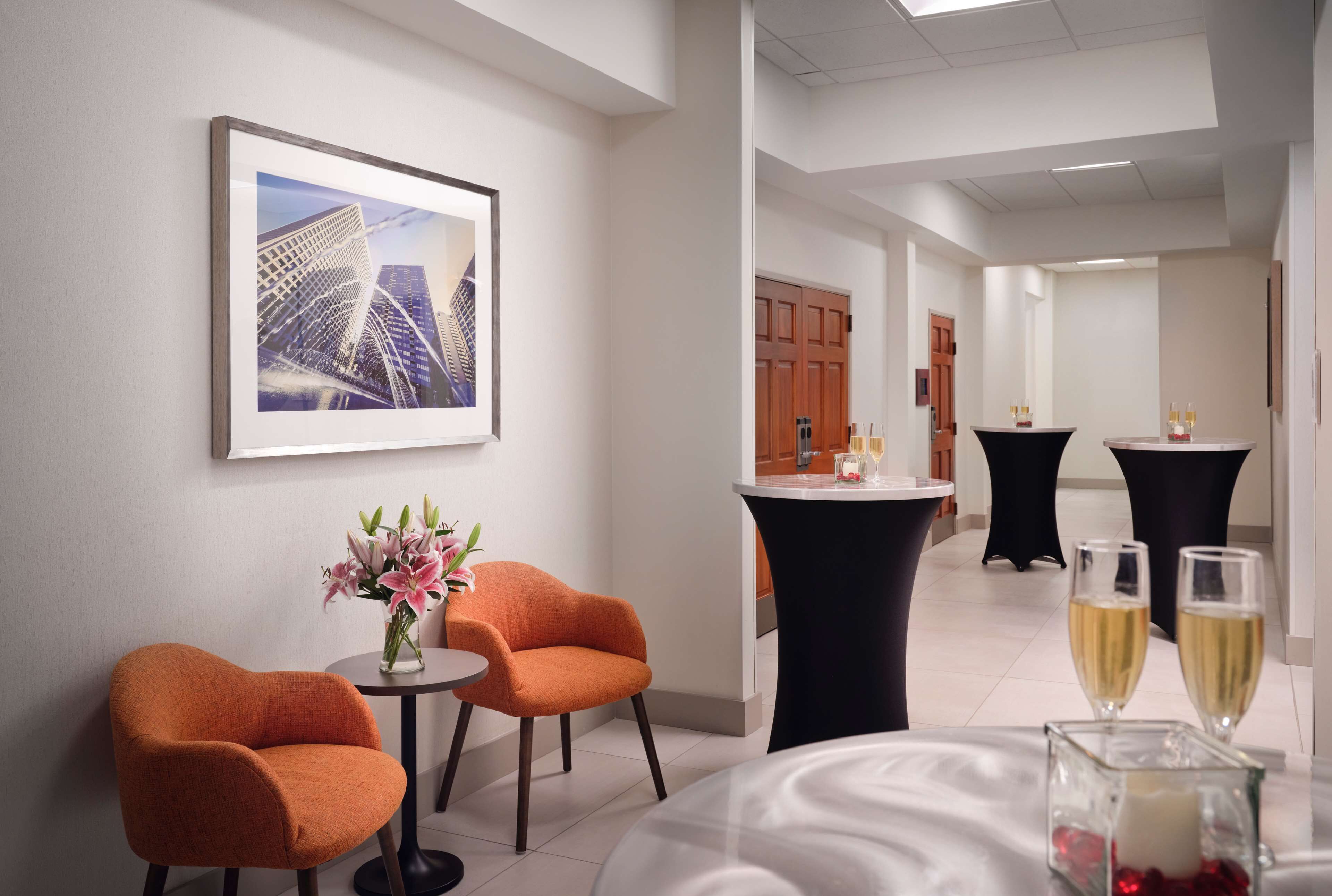 A hotel lobby with a modern design, featuring a framed cityscape artwork, orange chairs, a round table with a vase of flowers, and a bar area with two glasses of champagne.