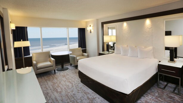 A hotel room with a large bed, a desk, a chair, a small table, and a view of the beach.