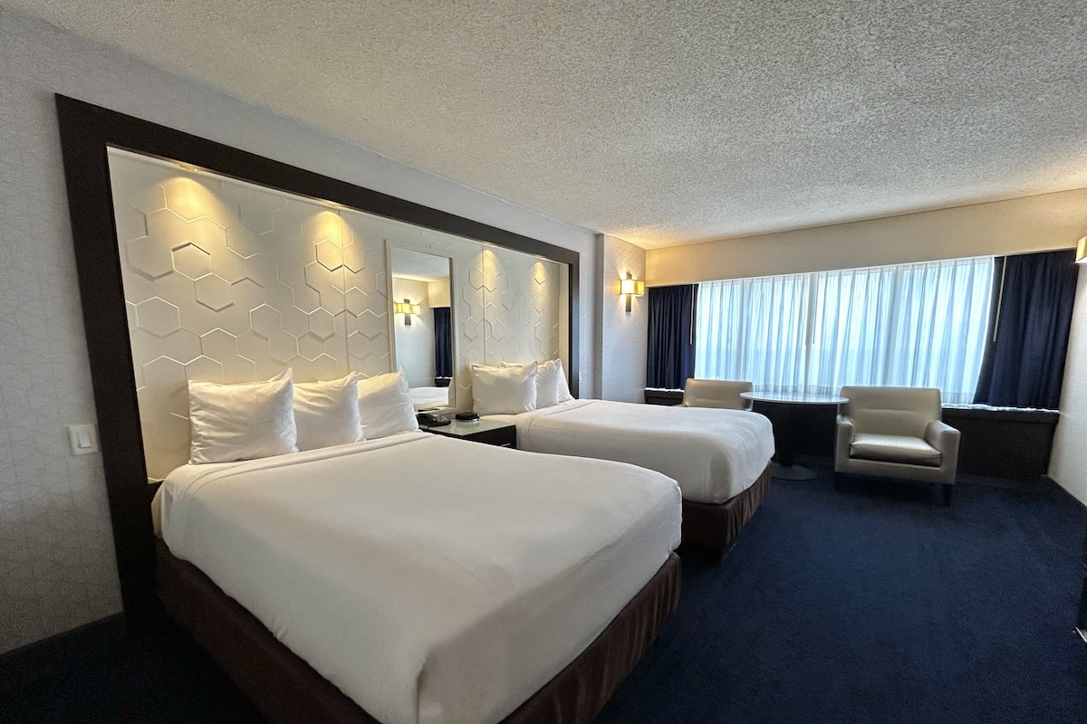 Une chambre d’hôtel avec deux lits, un bureau, une chaise et une grande fenêtre avec des rideaux.